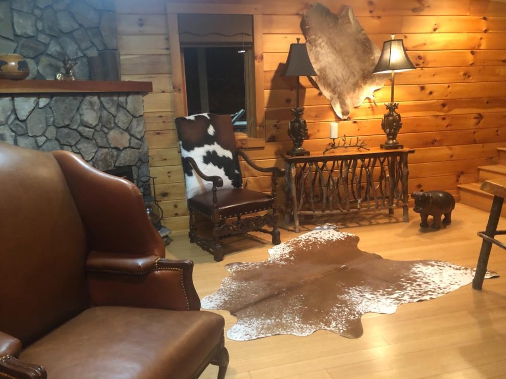Brown and white cowhide hung on a wood-paneled wall above a rustic console table, with a speckled cowhide rug on the floor