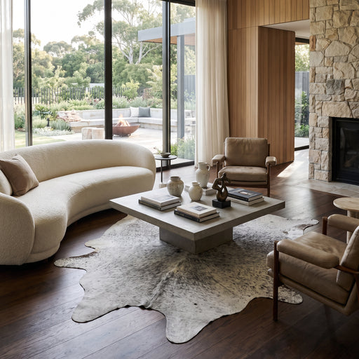 XXL tricolor speckled Brazilian cowhide rug styled in a modern, sunlit living room with a curved sofa and leather chairs.