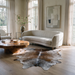 Brown salt and pepper cowhide rug styled on a light wood floor in a bright living room, beside a curved cream sofa and a wood coffee table