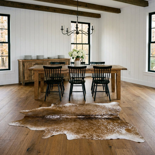 Brown salt and pepper cowhide rug on the oak floor of a modern farmhouse dining room with a rustic wood table and black Windsor chairs